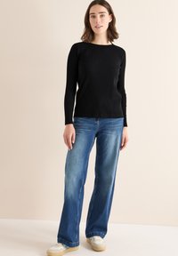 Zwarte geribbelde lange mouwen top gecombineerd met lichtblauwe wijde jeans en witte sneakers, tegen een neutrale achtergrond.