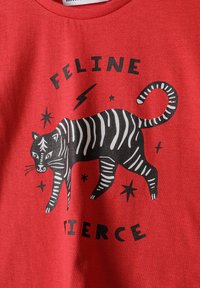 Crvena majica s crno-bijelim prugastim dizajnom crtanog mačka, uz natpis "FELINE FIERCE" i zvjezdaste akcentne detalje. Napravljena od mekog materijala.