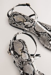 Zwart-witte sandalen met een slangenhuidpatroon, platte zolen, enkelbandjes en gouden gespsluitingen op een lichte achtergrond.