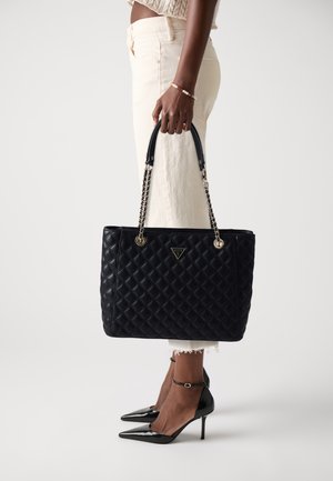 Schwarze gesteppte Tasche mit Kettenhenkeln, goldenen Akzenten und einem dreieckigen Logo. Tragen mit cremefarbenen Hosen und schwarzen Stilettos.