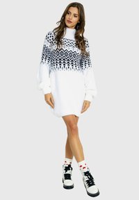 Biała sukienka swetrowa z czarnym geometrycznym wzorem u góry, długimi rękawami i wysokim kołnierzem. Połączona z czarno-białymi bucikami i skarpetkami z czerwonym akcentem.