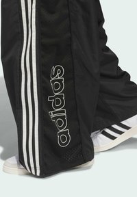 Čierne športové nohavice sú zdobené bielym trojpruhovým dizajnom a veľkým logom "adidas". Materiál je hladký s odvetranými panelmi pre lepšiu priedušnosť.