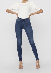 Top blanco de manga corta con mangas abullonadas, combinado con jeans ajustados de cintura alta de color azul oscuro. Tacones con tiras transparentes completan el atuendo.