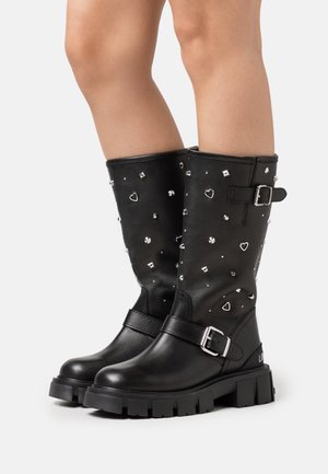 Zwarte laarzen tot halverwege de kuit met chunky zolen, zilveren gespen en decoratieve studs, waaronder hartjes en ronde vormen, op versleten benen.