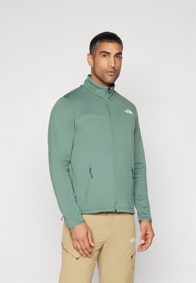 Giacca in pile verde con zip, colletto alto, due tasche anteriori con zip e una finitura in tessuto testurizzato. Presenta un logo sul petto.