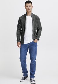 Cárdigan gris de punto con cremallera al frente, utilizado sobre una camiseta blanca, combinado con jeans de mezclilla azules y zapatillas negras con detalles blancos.