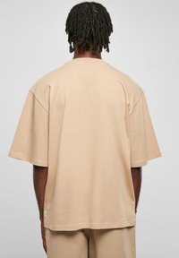 Tricou oversized bej cu mânecă scurtă, realizat dintr-un material moale. Prezintă o margine dreaptă și un eticheta mică la cusătura laterală.
