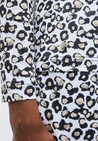 Κοντινή λήψη μανικιού και τσέπης ρούχου με λεοπάρ μοτίβο και μικρό ασημένιο έμβλημα σε σχήμα καρδιάς σε υφασμένο λευκό ύφασμα.