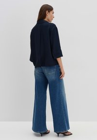 Camicia blu navy con texture, colletto e maniche arrotolate, abbinata a jeans a gamba larga di un leggero blu e sandali neri con tacco.