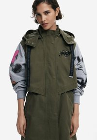Veste verte olive avec capuche, manches de sweat-shirt gris ornées de motifs de souris, rayures verticales et texte brodé sur le devant.