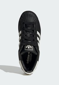 Czarne trampki z teksturowanym czarnym gumowym noskiem, białe trzy paski i miękki szary futrzany akcent wzdłuż boków. Czarne sznurowadła i logo.