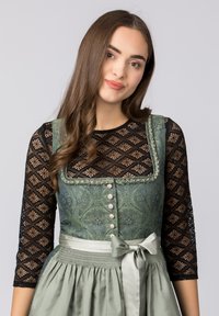 Grünes, gemustertes Dirndl mit schwarzen Spitzenärmeln, verziert mit Knöpfen und einer satinierten Schleife. Das strukturierte Material und die geraffte Taillenpartie heben das Design hervor.