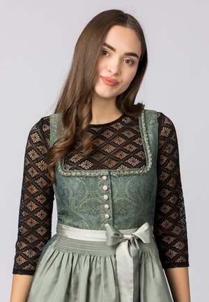 Grünes, gemustertes Dirndl mit schwarzen Spitzenärmeln, verziert mit Knöpfen und einer satinierten Schleife. Das strukturierte Material und die geraffte Taillenpartie heben das Design hervor.