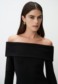 Abito nero a coste con spalle scoperte e maniche lunghe, caratterizzato da un design aderente. Orecchini a cerchio dorati completano il look. Texture liscia.