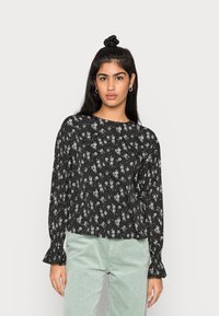 Blusa preta de manga longa com padrão floral, apresentando um decote redondo e mangas franzidas. Combinada com calças de corduroy verde claro.