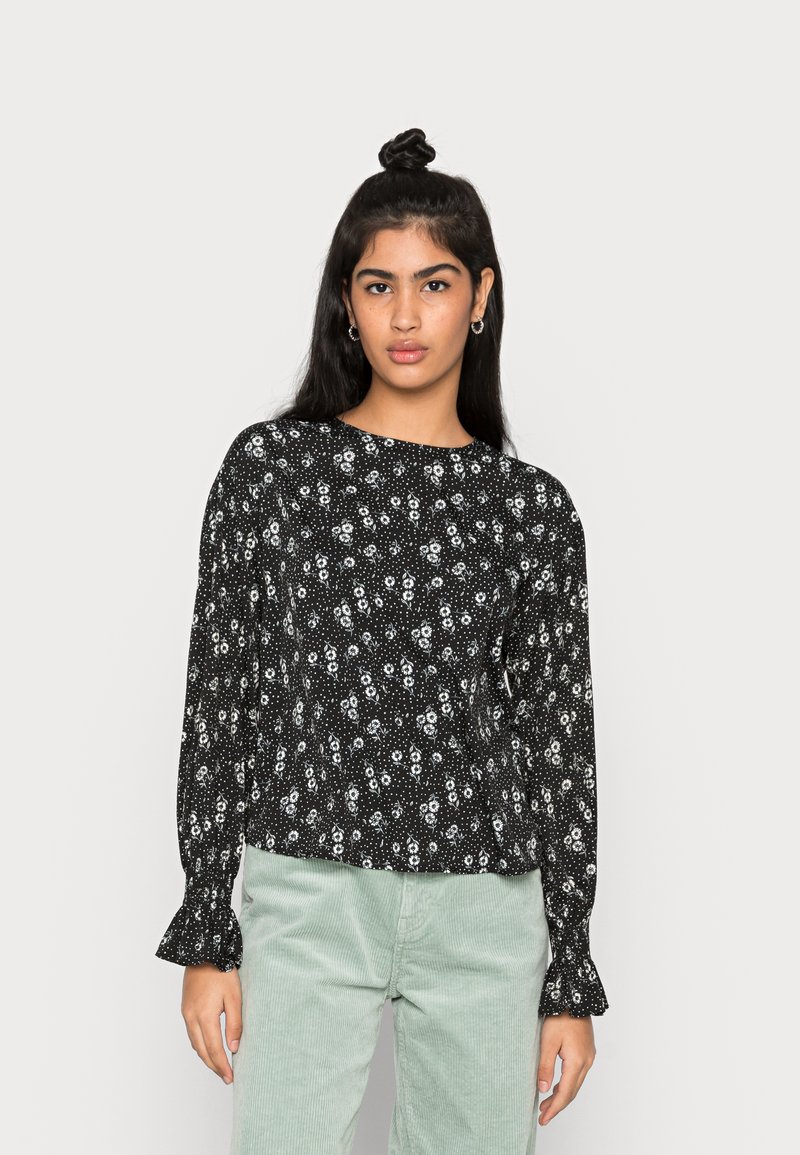 Blusa preta de manga longa com padrão floral, apresentando um decote redondo e mangas franzidas. Combinada com calças de corduroy verde claro.