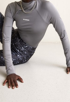Person wearing a gray long-sleeve top and patterned pants, sitting on the floor with hands supporting their body.