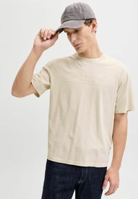 Beige Baumwoll-T-Shirt mit kurzen Ärmeln und Rundhalsausschnitt, versehen mit dezentem Logo-Text. Das Modell trägt dunkle Denim-Jeans und eine graue Mütze.