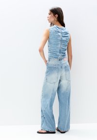 Prilagojen svetlo modri denim top z nabranimi detajli in zadrgo na hrbtu, v kombinaciji s širokimi usklajenimi kavbojkami z določenimi žepi in zbledelimi površinami.