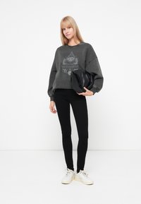 Grijze sweatshirt met een gedrukt oogontwerp, zwarte skinny jeans en witte sneakers. Houdt een zwarte tas vast met een ronde vorm.