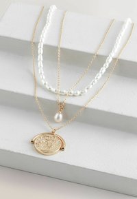 Collier en couches avec des accents en or et perles. Chaîne en or avec un pendentif rond texturé, associé à une suite de perles blanches.