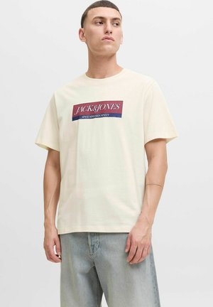 Camiseta de manga corta en color crema con un logo rectangular en burdeos y azul marino, confeccionada en suave tela de algodón, con un corte relajado.