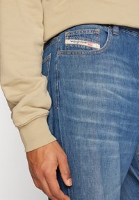 Jean en denim bleu légèrement délavé, avec une étiquette sur la poche arrière, et un pull de couleur kaki aux poignets côtelés. Gros plan sur une main posée sur le jean.