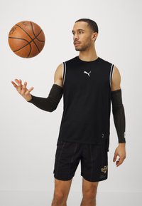 Maglietta sportiva senza maniche nera con strisce bianche, pantaloni corti neri con dettagli in rete e maniche per le braccia, che tiene una pallacanestro su uno sfondo neutro.
