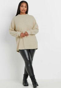 Beige ribbad stickad tröja med långa ärmar, tillsammans med blanka svarta leggings och svarta snörstövlar, placerad mot en vit bakgrund.