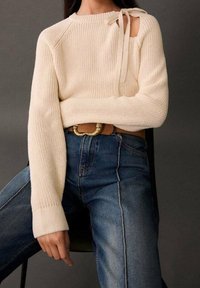 Kremowy sweter z dzianiny z ukośnym wycięciem i kokardą przy dekolcie, w połączeniu z niebieskimi dżinsami z wysokim stanem.