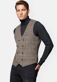 Brązowa kamizelka w kratkę z dekoltem w kształcie litery V, podwójnym zapięciem, czterema guzikami i dwiema bocznymi kieszeniami, noszona na czarnym golfie.