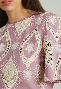 Lavendelfarbene Bluse mit aufwendiger cremefarbener Spitze, die über Blumendesigns und gewellte Akzente an Hals- und Ärmelöffnungen verfügt.