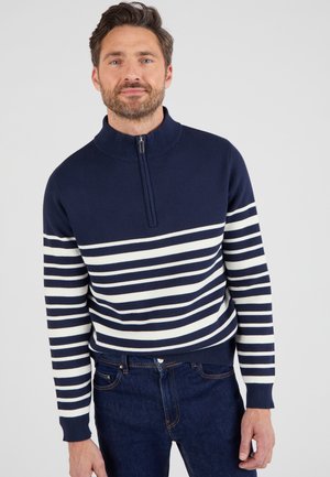 Jersey rayado en azul marino y blanco con un cuello alto y cierre de cremallera. Material suave, diseño ajustado, puños de canalé y dobladillo. Combinado con jeans oscuros.