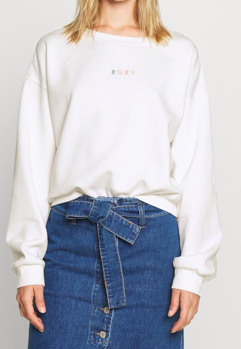 Femme portant un sweat-shirt blanc Roxy et une jupe en jean bleue taille haute nouée avec une ceinture devant.