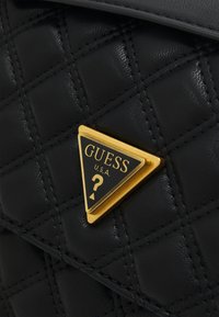 Juodas siuvinėtas dirbtinės odos rankinė su auksine trikampio formos logotipo plokštele, kurioje įspaustas užrašas "GUESS U.S.A." ir klaustuko ikona.