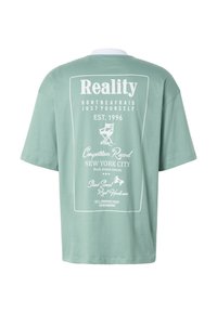 T-shirt oversize vert clair avec texte et graphismes imprimés en blanc, incluant « Reality », un fauteuil de réalisateur et plusieurs phrases faisant référence à New York City.