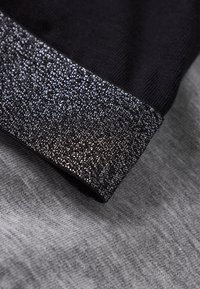 Μαύρο ύφασμα με μανσέτα που λάμπει με ασημί απόχρωση, με φόντο απαλό γκρι. Η μανσέτα παρουσιάζει υφή με αντανακλαστικές ιδιότ�ητες.