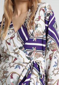 Abito a portafoglio con motivi floreali e a catena su un tessuto bianco. Strisce viola e blu navy accentuano il design. Fascia in vita.