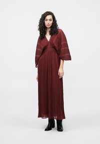 Burgunderfarget, lang plissett kjole med vide ermer, V-hals og innsvingt midje. Kombinert med svarte ankelstøvler. Glatt tekstur, flytende stoff.