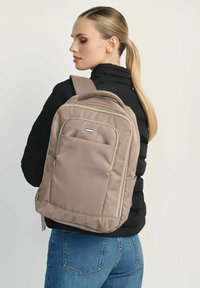 Beżowy plecak z wieloma przegrodami, zamkami z materiału i siateczkowym uchwytem na górze. Model nosi czarną kurtkę i niebieskie dżinsy.