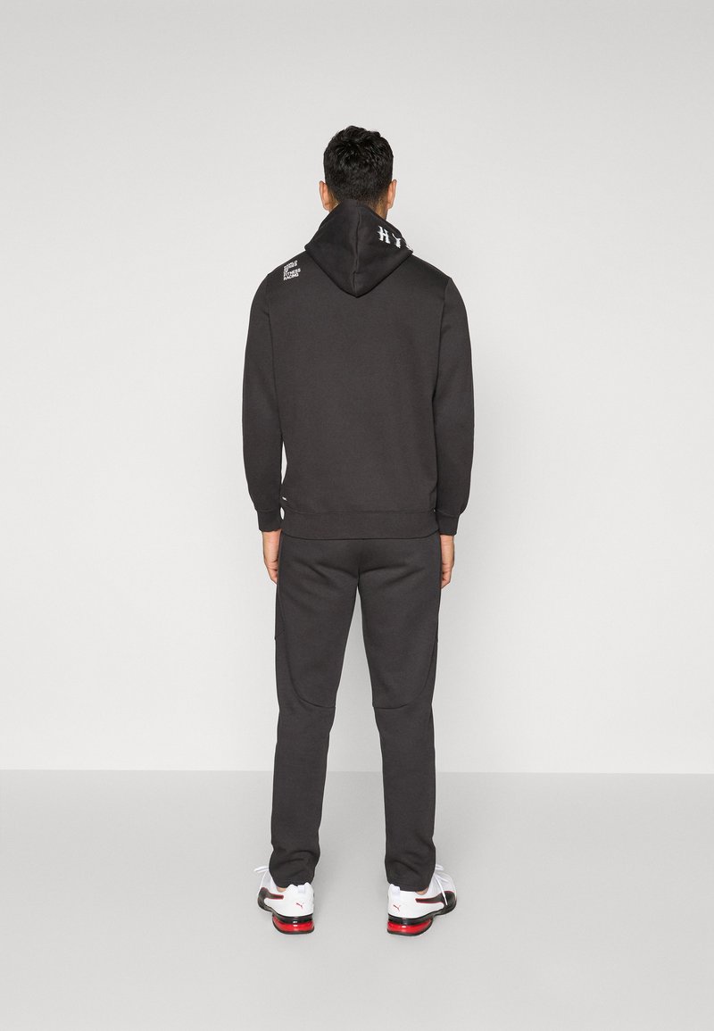 Sudadera negra con capucha, cordones y detalle de logotipo, combinada con pantalones de chándal a juego. El conjunto presenta un ajuste relajado y una textura suave.
