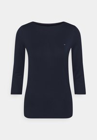 Tuum navy sininen paita, jossa on kolmivartiset hihat ja pieni punainen, valkoinen ja sininen logo vasemmassa rintapielessä valkoista taustaa vasten.