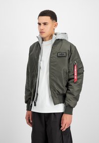 Chaqueta bomber verde oliva con forro de sudadera gris, puños de canalé y una etiqueta de tela roja. Incluye cierre delantero con cremallera y bolsillos laterales con cremallera.