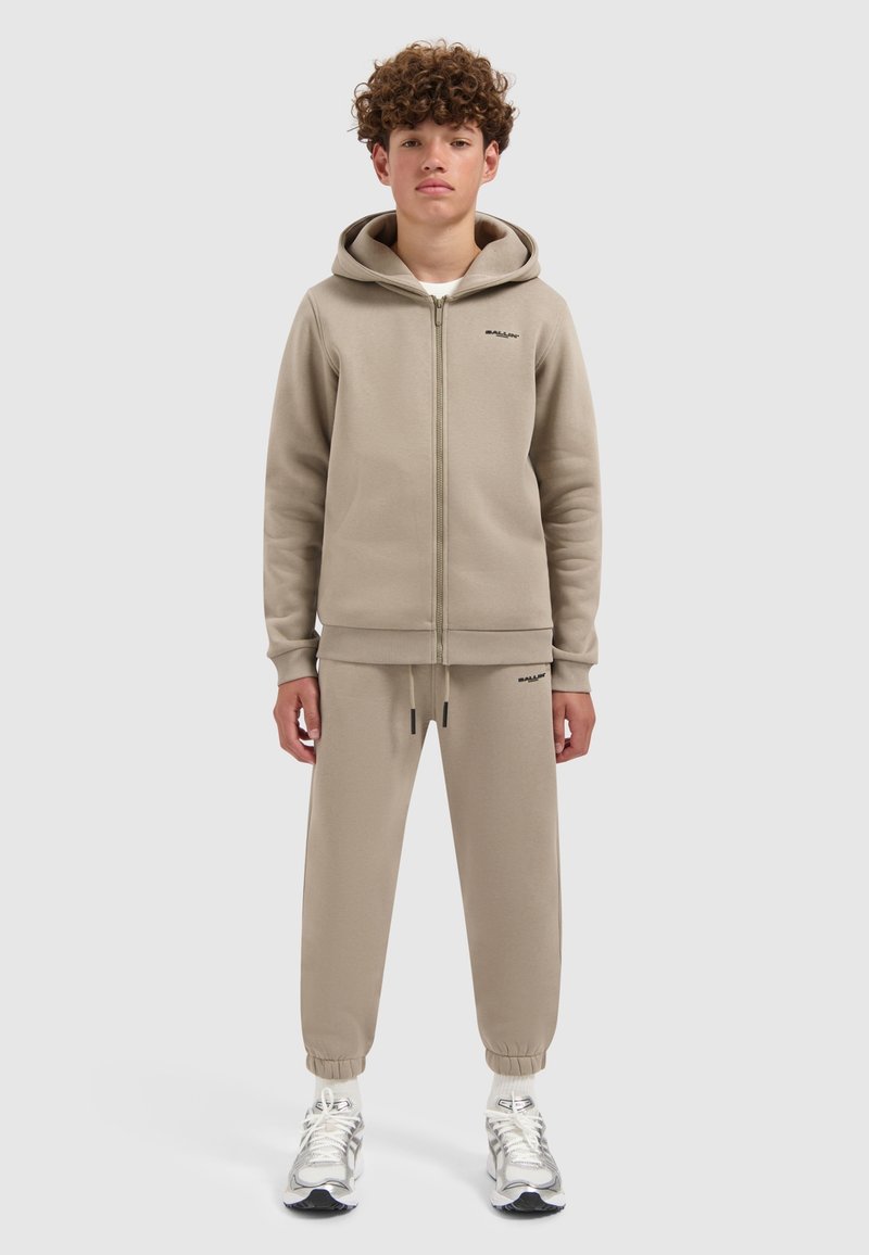 Tiener jongen staat rechtop en draagt een beige hoodie met ritssluiting en bijpassende jogpants met witte sneakers op een effen grijze achtergrond.