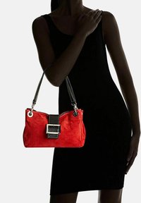 Punainen mokkanahkainen käsilaukku, jossa on musta nahkaremmi ja soljendetaili. Laukussa on hopeanväriset yksityiskohdat ja vetoketjukiinnitys.