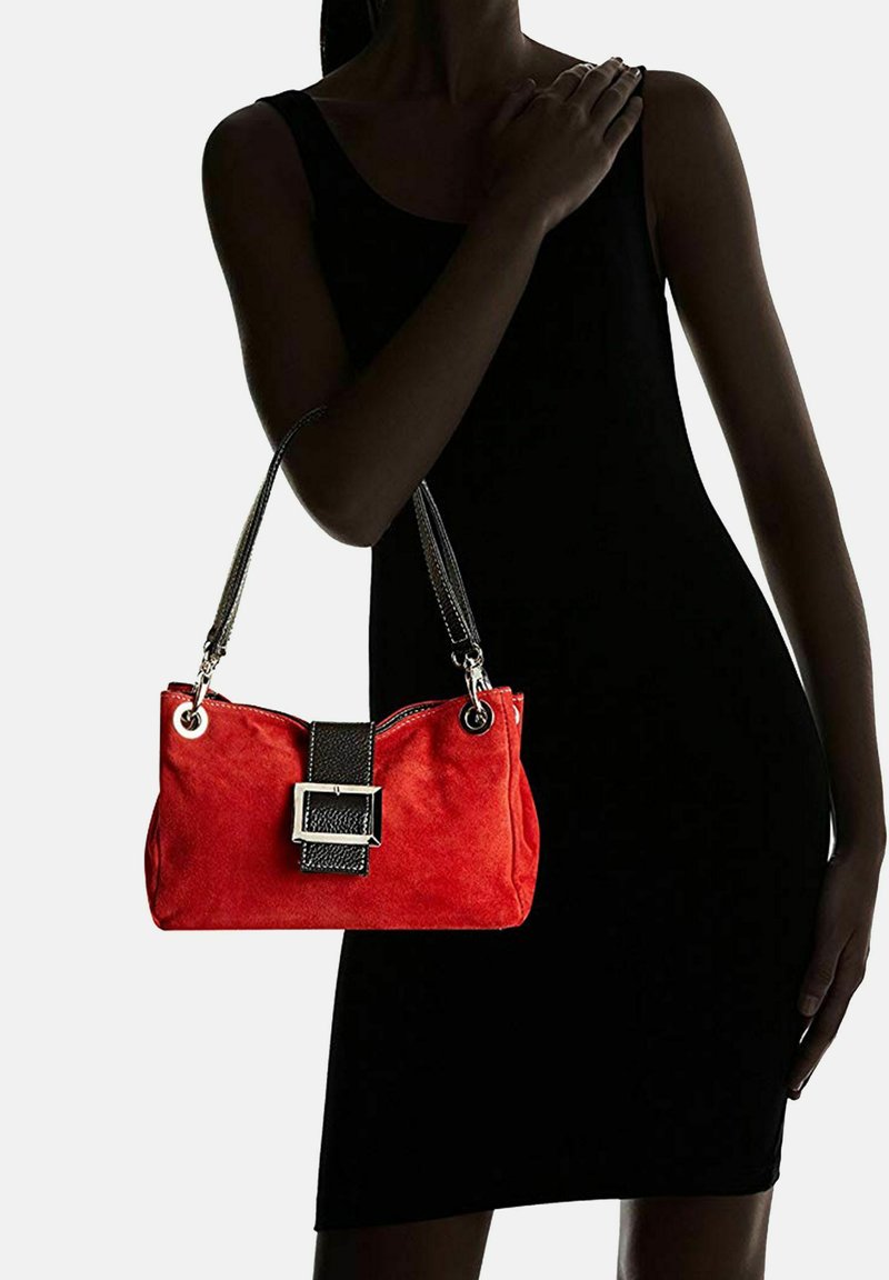Punainen mokkanahkainen käsilaukku, jossa on musta nahkaremmi ja soljendetaili. Laukussa on hopeanväriset yksityiskohdat ja vetoketjukiinnitys.