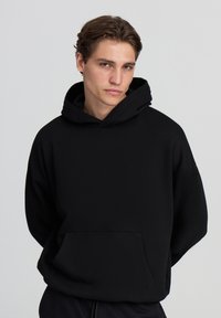 Felpa nera con cappuccio realizzata in morbido tessuto, dotata di tasca a canguro e vestibilità rilassata, con polsini e orlo a coste.