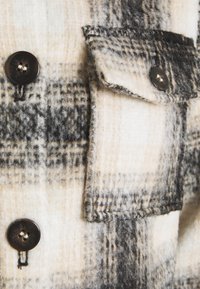 Camisa de flanela em padrão xadrez creme e preto com um bolso no peito, apresentando botões escuros e um acabamento suave e texturado.