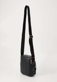 Borsa a tracolla nera dal design elegante, con un compartimento con zip e una tracolla regolabile su uno sfondo bianco uniforme.