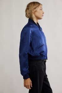 Modrá saténová bomber bunda s vysokým límcem, předním zipem a žebrovanými manžetami. Nahoře přes černé kalhoty s viditelnými bočními kapsami.